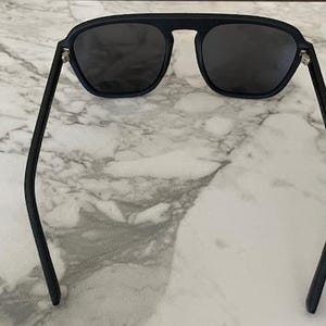 Puede incluir: Gafas de sol negras con un dise&ntilde;o de barra superior plana y lentes tintadas oscuras. La montura tiene un acabado mate con patillas finas y curvadas, y bisagras de metal plateado que conectan el frente de la montura con los brazos.