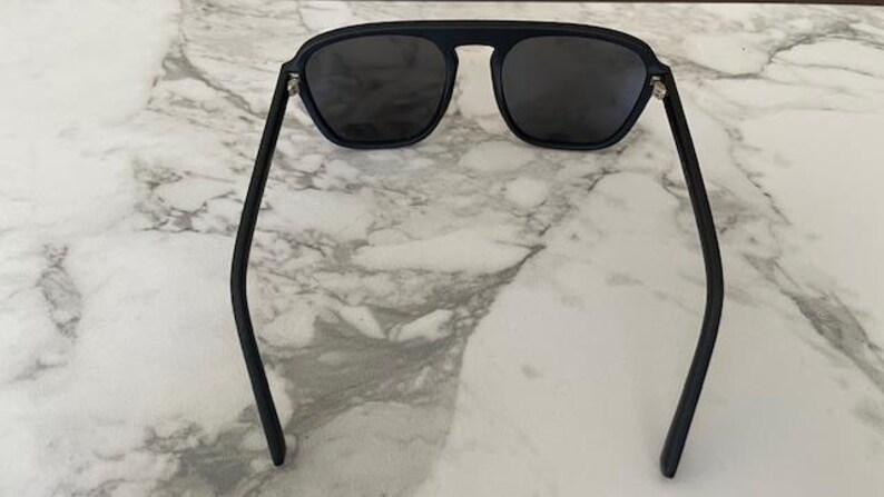 Puede incluir: Gafas de sol negras con un dise&ntilde;o de barra superior plana y lentes tintadas oscuras. La montura tiene un acabado mate con patillas finas y curvadas, y bisagras de metal plateado que conectan el frente de la montura con los brazos.