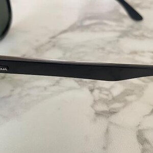 Puede incluir: Gafas de sol Ray-Ban negras con lentes oscuras. Las gafas tienen una montura negra brillante y el logotipo de Ray-Ban es visible en la patilla. Las gafas descansan sobre una superficie de m&aacute;rmol blanco y gris.