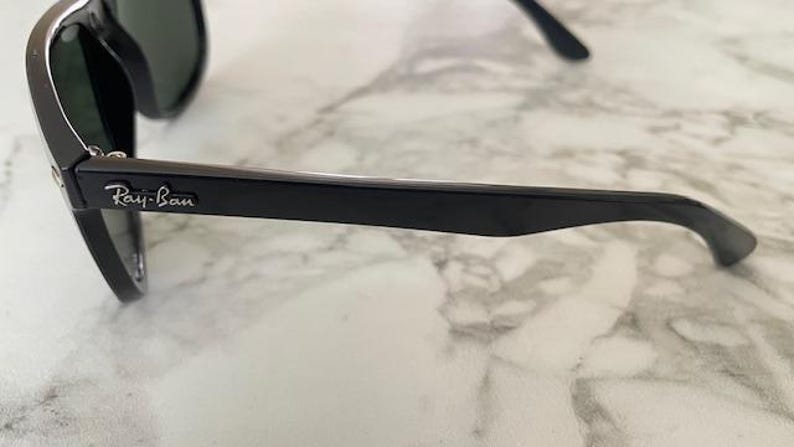 Puede incluir: Gafas de sol Ray-Ban negras con lentes oscuras. Las gafas tienen una montura negra brillante y el logotipo de Ray-Ban es visible en la patilla. Las gafas descansan sobre una superficie de m&aacute;rmol blanco y gris.