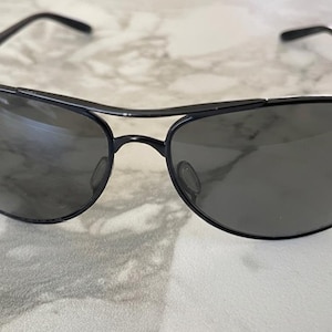May include: Black aviator sunglasses with a double bridge and dark lenses. The sunglasses have a sleek, modern design with a metal frame. The lenses are a dark gray color, and the overall look is stylish and classic.