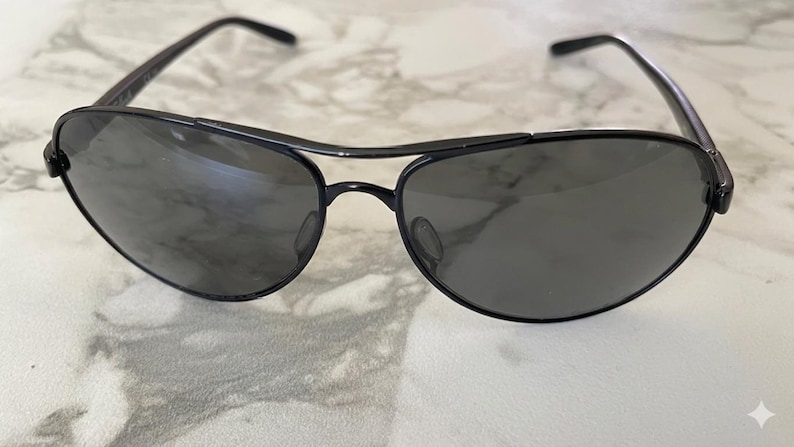 May include: Black aviator sunglasses with a double bridge and dark lenses. The sunglasses have a sleek, modern design with a metal frame. The lenses are a dark gray color, and the overall look is stylish and classic.