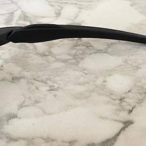Peut inclure: Gros plan sur une monture de lunettes de soleil de sport noire avec une branche incurv&eacute;e. Le verre est de couleur vert clair. Les lunettes de soleil reposent sur une surface en marbre blanc et gris.