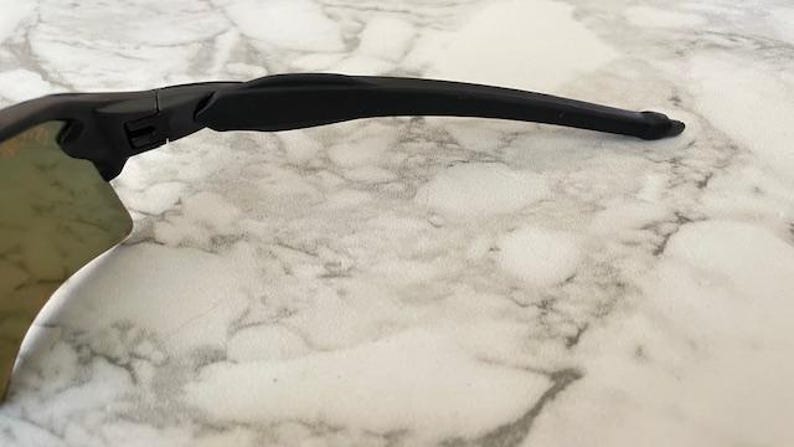 Peut inclure: Gros plan sur une monture de lunettes de soleil de sport noire avec une branche incurv&eacute;e. Le verre est de couleur vert clair. Les lunettes de soleil reposent sur une surface en marbre blanc et gris.