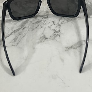 May include: Black sunglasses with dark lenses. The sunglasses have a rectangular frame and sleek, curved arms. The lenses are a dark gray color, and the frame is a glossy black. The sunglasses are set against a white and gray marble background.