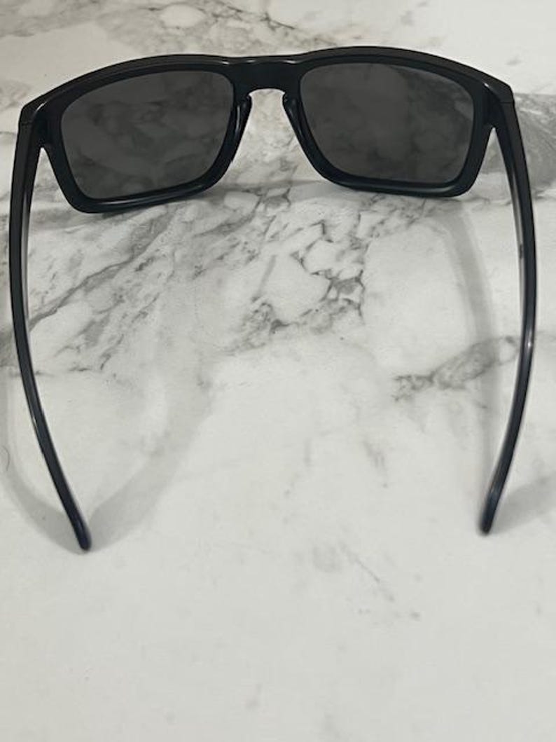 May include: Black sunglasses with dark lenses. The sunglasses have a rectangular frame and sleek, curved arms. The lenses are a dark gray color, and the frame is a glossy black. The sunglasses are set against a white and gray marble background.