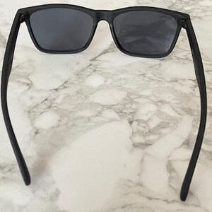 Pu&ograve; includere: Occhiali da sole neri con lenti scure. Gli occhiali hanno una montatura rettangolare e aste dritte. L'immagine &egrave; ripresa dall'alto, mostrando gli occhiali da sole su uno sfondo di marmo.