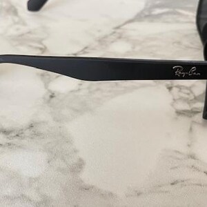 Pu&ograve; includere: Occhiali da sole neri con finitura opaca. Il logo Ray-Ban &egrave; visibile sull'asta. Gli occhiali sono appoggiati su una superficie marmorizzata.
