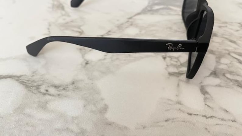 Pu&ograve; includere: Occhiali da sole neri con finitura opaca. Il logo Ray-Ban &egrave; visibile sull'asta. Gli occhiali sono appoggiati su una superficie marmorizzata.