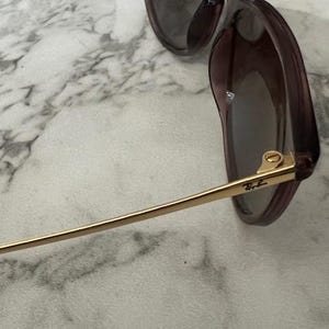 May include: Close-up of sunglasses with a dark purple frame and tinted lenses. The temples are gold-colored with the Ray-Ban logo. The sunglasses are resting on a white and gray marble surface.