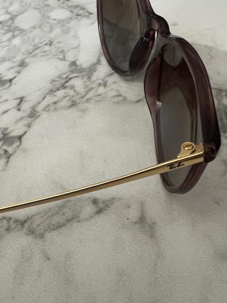 May include: Close-up of sunglasses with a dark purple frame and tinted lenses. The temples are gold-colored with the Ray-Ban logo. The sunglasses are resting on a white and gray marble surface.