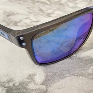 May include: Dark gray sunglasses with blue and purple reflective lenses. The sunglasses have a rectangular frame and a small logo on the temple. The lenses reflect the surrounding light, creating a vibrant color effect.