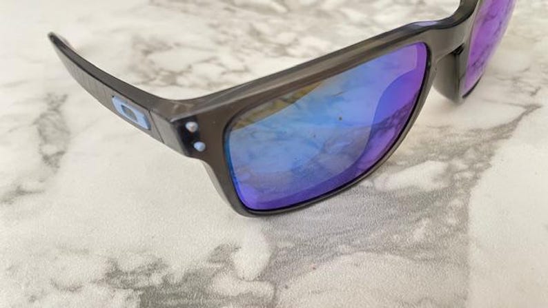May include: Dark gray sunglasses with blue and purple reflective lenses. The sunglasses have a rectangular frame and a small logo on the temple. The lenses reflect the surrounding light, creating a vibrant color effect.
