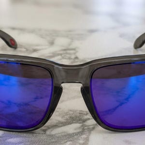 May include: Gray sunglasses with square frames and blue mirrored lenses. The temples are dark gray with a small red accent. The lenses reflect a bright blue hue, and the frames have small, light-colored accents.