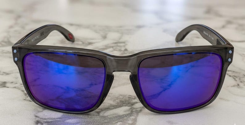 May include: Gray sunglasses with square frames and blue mirrored lenses. The temples are dark gray with a small red accent. The lenses reflect a bright blue hue, and the frames have small, light-colored accents.