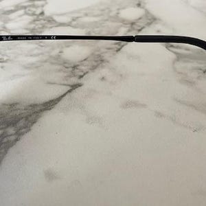 May include: Black sunglasses with gradient blue lenses. The frame is black, with the text "MADE IN ITALY" and a logo on the temple. The sunglasses are resting on a white and gray marble surface.