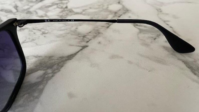 May include: Black sunglasses with gradient blue lenses. The frame is black, with the text "MADE IN ITALY" and a logo on the temple. The sunglasses are resting on a white and gray marble surface.