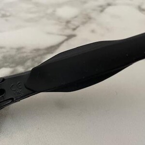 Puede incluir: Primer plano de gafas de sol negras con acabado mate. La montura tiene un dise&ntilde;o elegante y moderno con una patilla curva. Las lentes tienen un tinte degradado y el &aacute;rea de la bisagra muestra una marca CE.