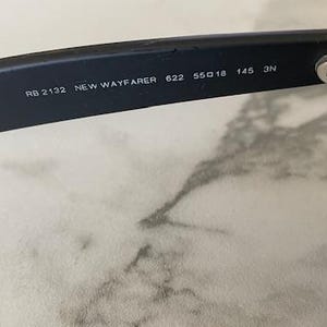 May include: Close-up of black sunglasses with the text "RB 2132 NEW WAYFARER 622 55O18 145 3N" printed on the arm. The sunglasses have a classic design with dark lenses and a black frame.