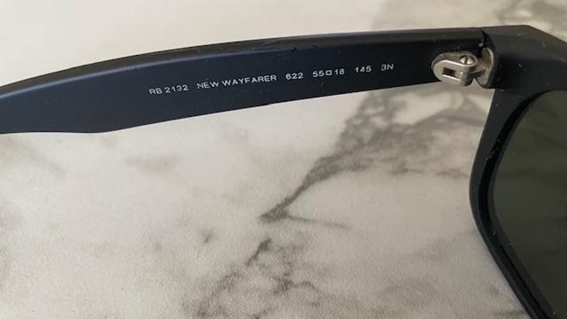 May include: Close-up of black sunglasses with the text "RB 2132 NEW WAYFARER 622 55O18 145 3N" printed on the arm. The sunglasses have a classic design with dark lenses and a black frame.