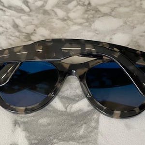 Puede incluir: Gafas de sol con montura de carey y lentes azules. Las gafas de sol tienen un dise&ntilde;o cl&aacute;sico con una barra superior curvada y patillas gruesas. El logotipo de Ray-Ban es visible.