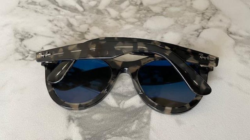 Puede incluir: Gafas de sol con montura de carey y lentes azules. Las gafas de sol tienen un dise&ntilde;o cl&aacute;sico con una barra superior curvada y patillas gruesas. El logotipo de Ray-Ban es visible.