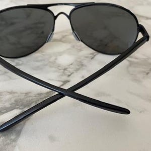 May include: Black aviator sunglasses with dark lenses. The sunglasses have a thin, black metal frame and a double bridge. The arms of the sunglasses are crossed, resting on a marble surface.