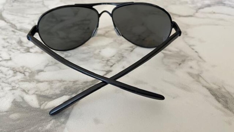 May include: Black aviator sunglasses with dark lenses. The sunglasses have a thin, black metal frame and a double bridge. The arms of the sunglasses are crossed, resting on a marble surface.