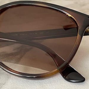 May include: Brown tortoiseshell sunglasses with brown tinted lenses. The frames have a rounded shape and a glossy finish. The arms of the sunglasses are dark brown and have a slight curve. The lenses are a medium brown color.