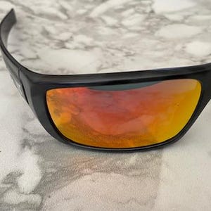 Puede incluir: Gafas de sol negras con lentes espejadas naranjas y amarillas. Las gafas de sol tienen un dise&ntilde;o elegante y curvado y est&aacute;n sobre un fondo de m&aacute;rmol. Las lentes reflejan el entorno.