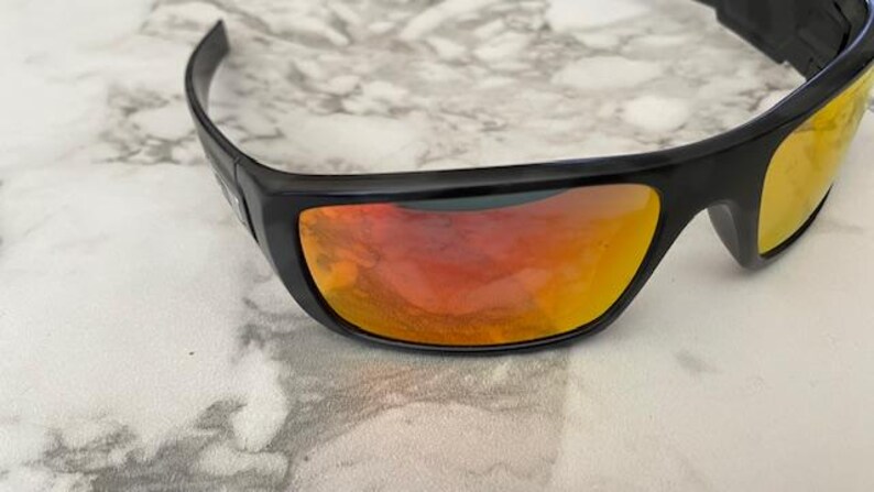Puede incluir: Gafas de sol negras con lentes espejadas naranjas y amarillas. Las gafas de sol tienen un dise&ntilde;o elegante y curvado y est&aacute;n sobre un fondo de m&aacute;rmol. Las lentes reflejan el entorno.