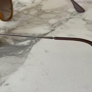 May include: Sunglasses with brown frames and lenses. The glasses have a silver metal arm that connects to a brown earpiece. The lenses are a light brown color. The sunglasses are on a white and gray marble surface.
