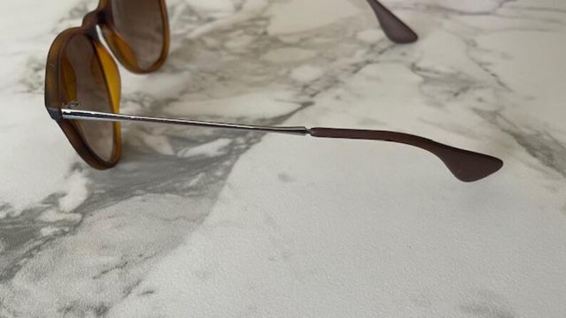 May include: Sunglasses with brown frames and lenses. The glasses have a silver metal arm that connects to a brown earpiece. The lenses are a light brown color. The sunglasses are on a white and gray marble surface.
