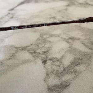 K&ouml;nnte beinhalten: Nahaufnahme des B&uuml;gels einer Ray-Ban Sonnenbrille. Der Rahmen ist dunkelbraun mit dem Text "MADE IN ITALY" und dem Ray-Ban-Logo. Der B&uuml;gel hat eine braune, gummierte Spitze.