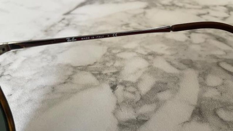 K&ouml;nnte beinhalten: Nahaufnahme des B&uuml;gels einer Ray-Ban Sonnenbrille. Der Rahmen ist dunkelbraun mit dem Text "MADE IN ITALY" und dem Ray-Ban-Logo. Der B&uuml;gel hat eine braune, gummierte Spitze.