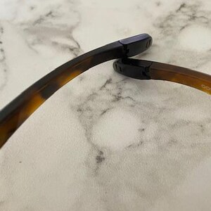 Op de afbeelding: Close-up van de poot van een bril. Het montuur heeft een schildpadpatroon, dat overgaat van donkerbruin naar een lichtere tint. De uiteinden zijn zwart. De tekst "OO9141-0354 44 17 132" is zichtbaar.