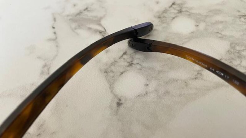 Op de afbeelding: Close-up van de poot van een bril. Het montuur heeft een schildpadpatroon, dat overgaat van donkerbruin naar een lichtere tint. De uiteinden zijn zwart. De tekst "OO9141-0354 44 17 132" is zichtbaar.