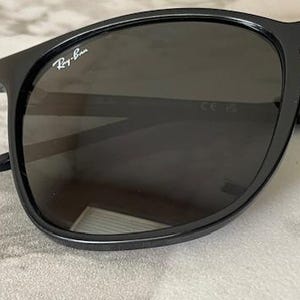 Puede incluir: Gafas de sol negras con lentes oscuras. Las monturas son brillantes y presentan el logotipo de Ray-Ban en blanco en la esquina superior izquierda. Las gafas de sol tienen una forma cl&aacute;sica y redondeada, sobre un fondo neutro.