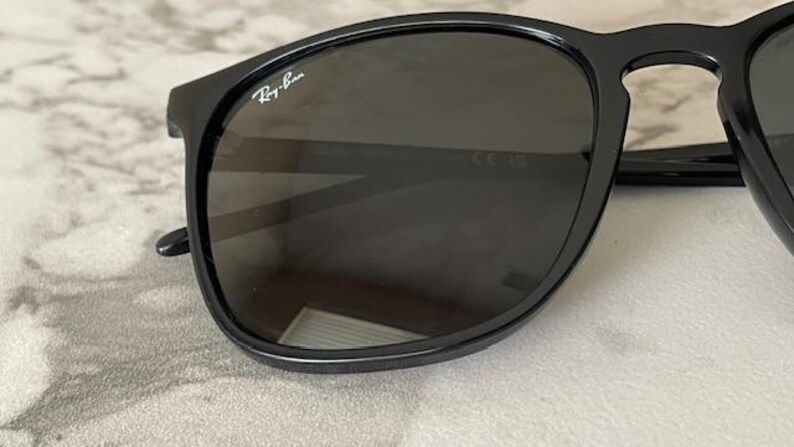Puede incluir: Gafas de sol negras con lentes oscuras. Las monturas son brillantes y presentan el logotipo de Ray-Ban en blanco en la esquina superior izquierda. Las gafas de sol tienen una forma cl&aacute;sica y redondeada, sobre un fondo neutro.