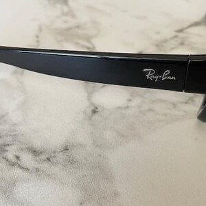 Puede incluir: Gafas de sol negras con el logotipo de Ray-Ban en la patilla. Las gafas de sol tienen un dise&ntilde;o elegante y moderno con un acabado brillante. Las monturas son gruesas y las lentes oscuras, ofreciendo protecci&oacute;n solar.