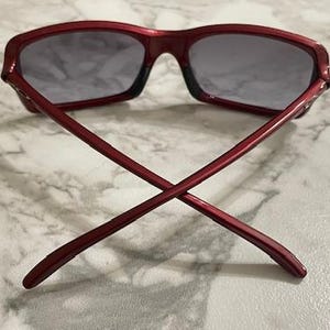 May include: Red sunglasses with dark lenses, viewed from the back with the arms crossed. The frame is a solid, glossy red. The lenses are a dark gray color. The sunglasses are resting on a light-colored, marbled surface.