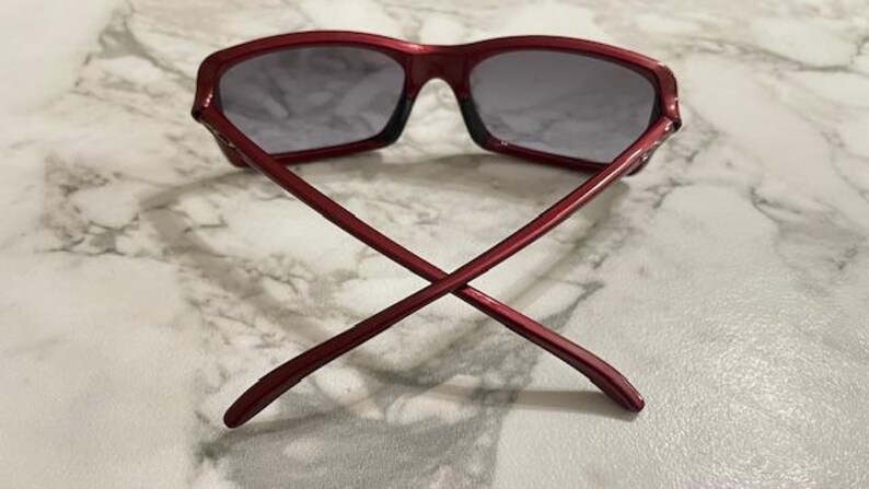 May include: Red sunglasses with dark lenses, viewed from the back with the arms crossed. The frame is a solid, glossy red. The lenses are a dark gray color. The sunglasses are resting on a light-colored, marbled surface.