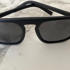 Puede incluir: Gafas de sol en negro mate con una barra superior gruesa y angular y lentes oscuras. La montura presenta un dise&ntilde;o s&oacute;lido y estructurado con un puente prominente que conecta los dos cristales cuadrados. Las varillas son finas y rectas.