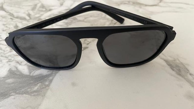 Puede incluir: Gafas de sol en negro mate con una barra superior gruesa y angular y lentes oscuras. La montura presenta un dise&ntilde;o s&oacute;lido y estructurado con un puente prominente que conecta los dos cristales cuadrados. Las varillas son finas y rectas.