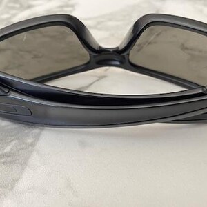 May include: Black sunglasses with a curved frame and grey lenses. The sunglasses have a sleek, modern design. The arms of the sunglasses have a gold emblem. The sunglasses are resting on a white surface.