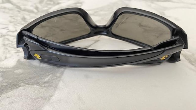 May include: Black sunglasses with a curved frame and grey lenses. The sunglasses have a sleek, modern design. The arms of the sunglasses have a gold emblem. The sunglasses are resting on a white surface.