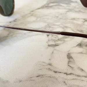 K&ouml;nnte beinhalten: Sonnenbrille mit braunem Gestell und gr&uuml;nen Gl&auml;sern. Die B&uuml;gel sind silber und braun, mit einem geschwungenen Design f&uuml;r einen bequemen Sitz. Die Brille liegt auf einem Marmorhintergrund.