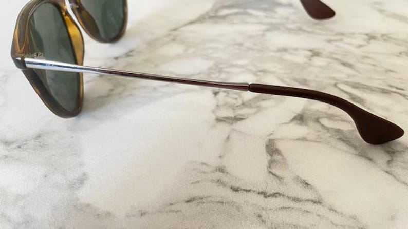 K&ouml;nnte beinhalten: Sonnenbrille mit braunem Gestell und gr&uuml;nen Gl&auml;sern. Die B&uuml;gel sind silber und braun, mit einem geschwungenen Design f&uuml;r einen bequemen Sitz. Die Brille liegt auf einem Marmorhintergrund.