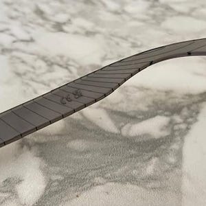 May include: Close-up of a gray eyeglass arm with a textured design. The arm has a red accent at the end and a small marking near the middle. The arm is curved and appears to be made of a durable material.