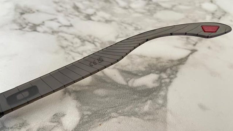 May include: Close-up of a gray eyeglass arm with a textured design. The arm has a red accent at the end and a small marking near the middle. The arm is curved and appears to be made of a durable material.